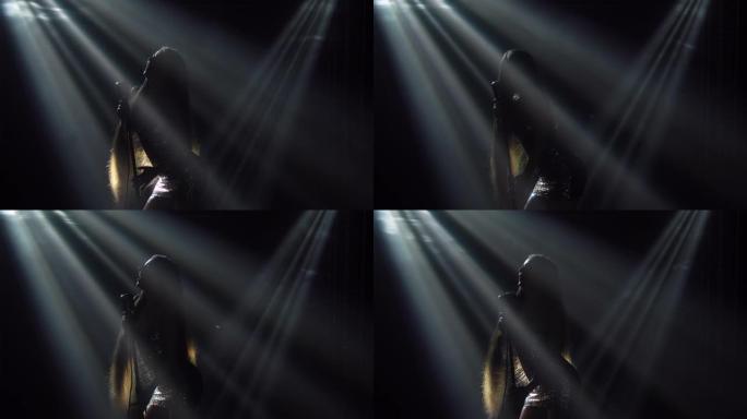 一个美丽的女人在聚光灯下黑暗的舞台上对着老式麦克风唱歌的肖像。这位年轻的歌手动情地唱着这首歌。展示商