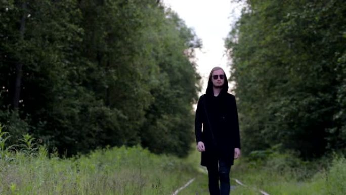 一个穿着黑色衣服的英俊年轻人在绿色森林的背景下沿着古老的铁轨行走。电影或剪辑的情节