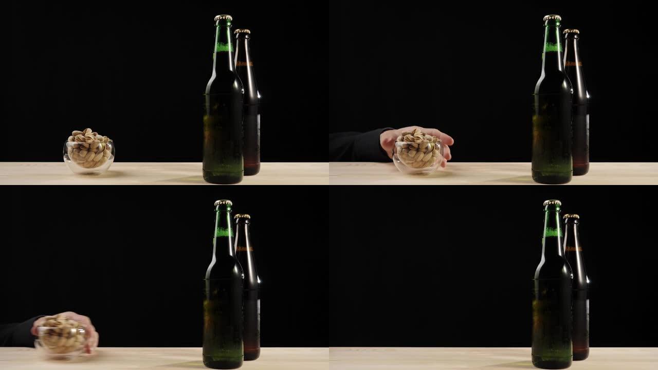 新鲜啤酒。在黑色背景下的木制桌子上，用手滑出开心果碟，靠近绿色和棕色的瓶子，上面放着美味的工艺啤酒。