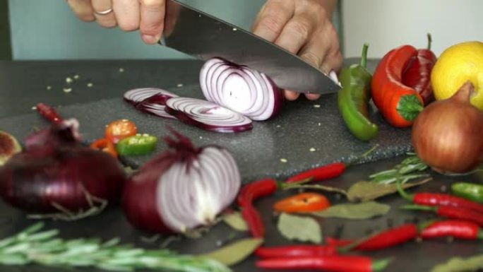 在蔬菜，香料和调味料的背景下，手在砧板上切红洋葱。厨师用蔬菜和香料准备菜