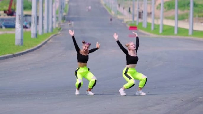 两个穿着运动服的年轻女孩正在跳嘻哈，在宽阔的街道上表演现代青年舞蹈的壮观元素。慢动作