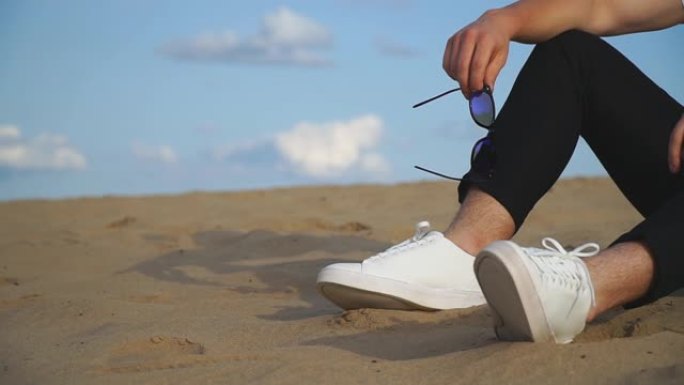 穿着时髦衣服的面目全非的家伙坐在沙滩上。手持太阳镜的男人在海岸放松。享受户外娱乐的成年男性或商人。成