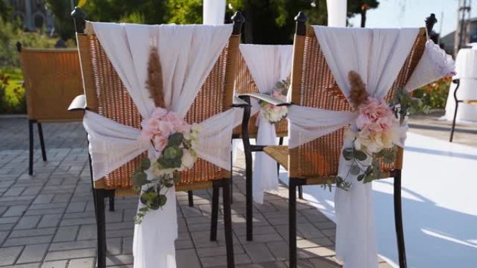 阳光明媚的夏日用鲜花装饰的婚礼场所。椅子上的节日花卉装饰。背景上白色纺织拱门上有球茎花环的玫瑰和羽毛