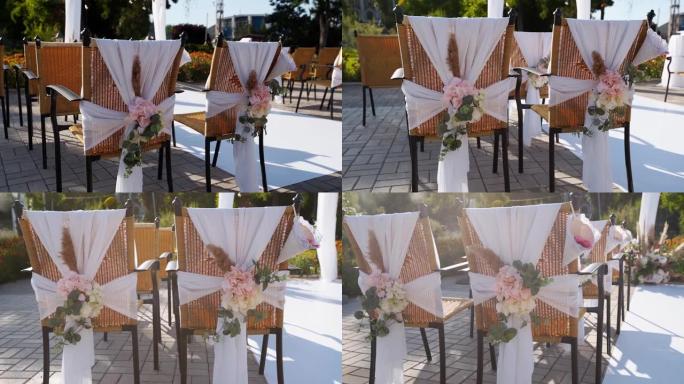 阳光明媚的夏日用鲜花装饰的婚礼场所。椅子上的节日花卉装饰。背景上白色纺织拱门上有球茎花环的玫瑰和羽毛