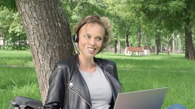 坐在城市公园时，带着笔记本电脑和耳机进行在线对话的女人