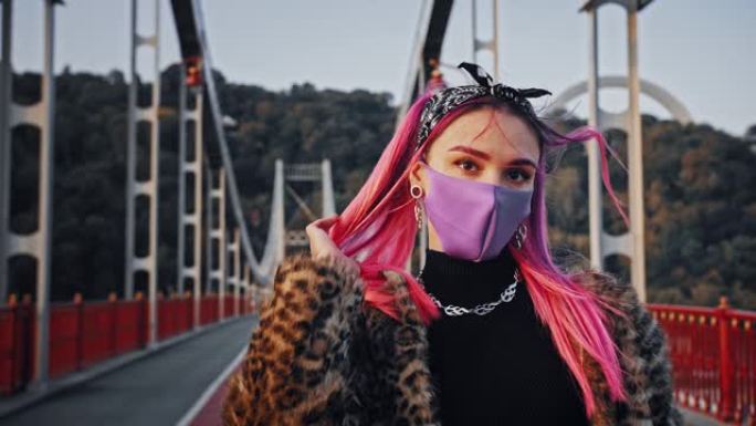 穿着非正式衣服和防护面具的年轻时髦女性正站在桥上拉直她的粉红色头发
