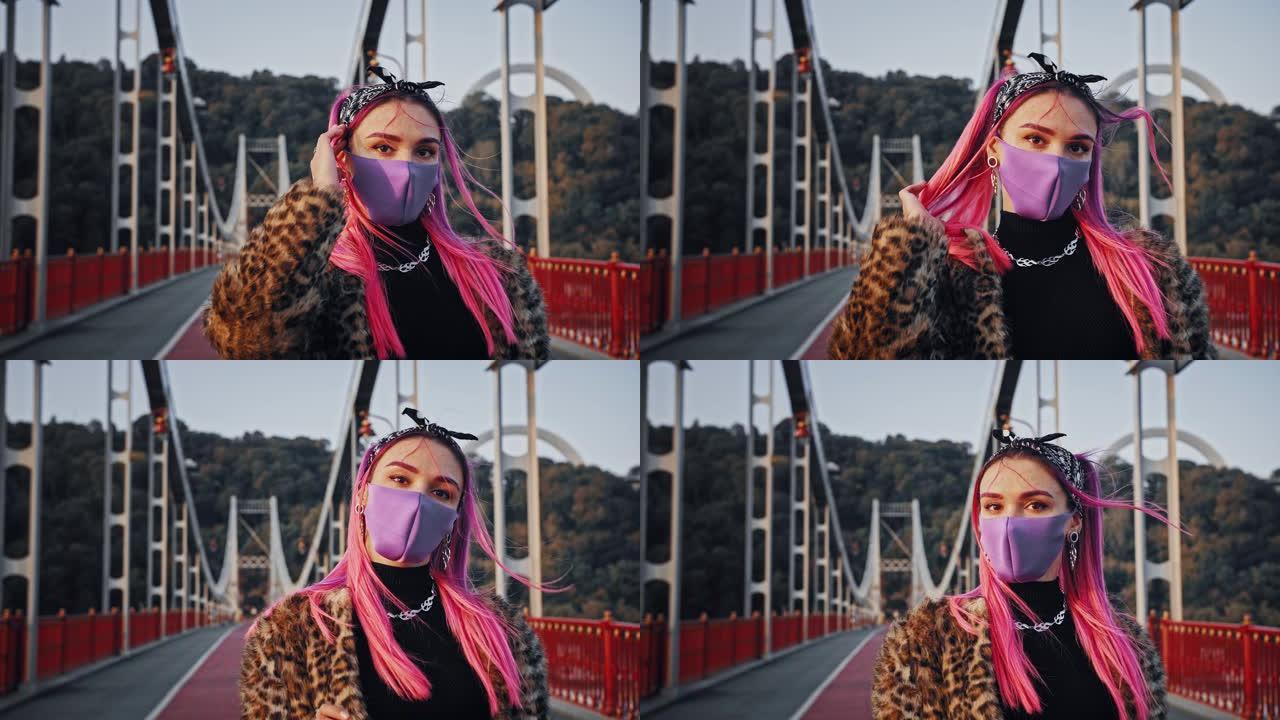 穿着非正式衣服和防护面具的年轻时髦女性正站在桥上拉直她的粉红色头发