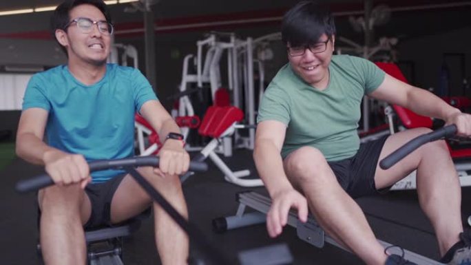 两名亚洲男子在互相取笑之前在划船机上工作，喜剧健身房概念