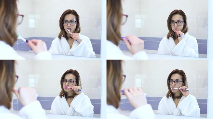 一个戴眼镜的年轻女子刷牙。一个穿白大褂的女人开始刷牙。透过镜子观看