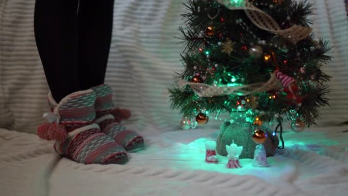 女人穿着温暖的冬季拖鞋在圣诞树下跳舞，装饰着五颜六色的花环，闪闪发光，照亮了房间。寒假和节日气氛