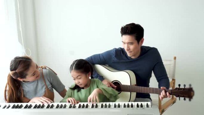 快乐的亚洲小女儿和妈妈一起弹钢琴，爸爸在家弹吉他，妈妈教女儿弹钢琴，他们弹唱歌曲。他们玩得很开心。