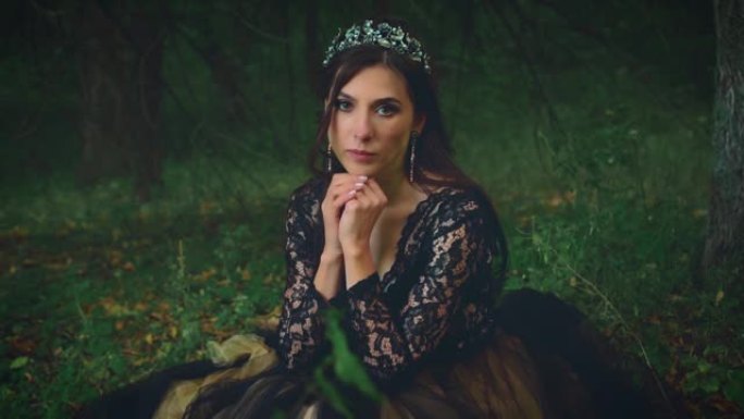 穿着黑色连衣裙的美女哥特式女王坐在黑暗的森林里，很悲伤。