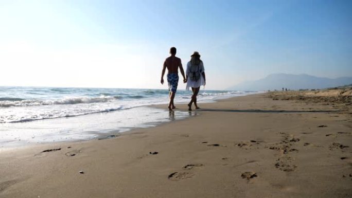 情侣在度假胜地休闲时牵手走在海边。男人和女人在海滩上漫步，享受暑假。年轻一对的蜜月。爱和幸福的概念。