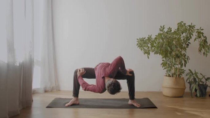 柔韧的男人做瑜伽室内阳光房背景慢动作。瑜伽大师站在平衡体式伸展腿部肌肉复制文本空间。健康生活方式身体