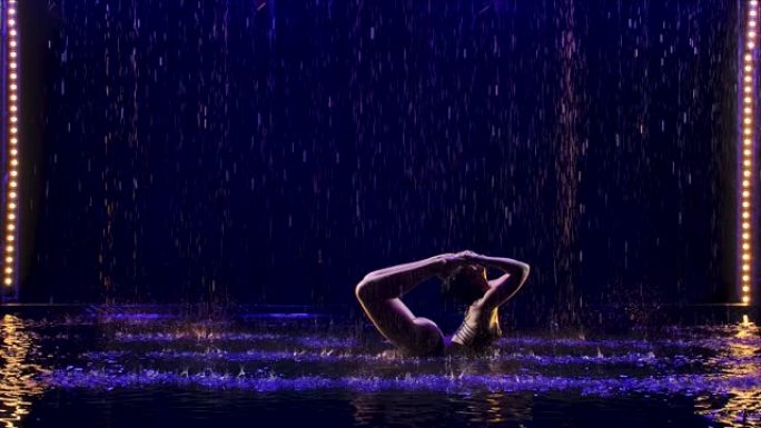一名年轻女运动员在雨中黑暗的工作室里表演的弓姿势。Studio light在水面和身体上产生微光。健