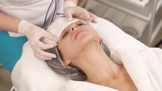 专业治疗师用白色餐巾纸清洁女性客户的脸部和颈部的特写镜头。美容和保健概念