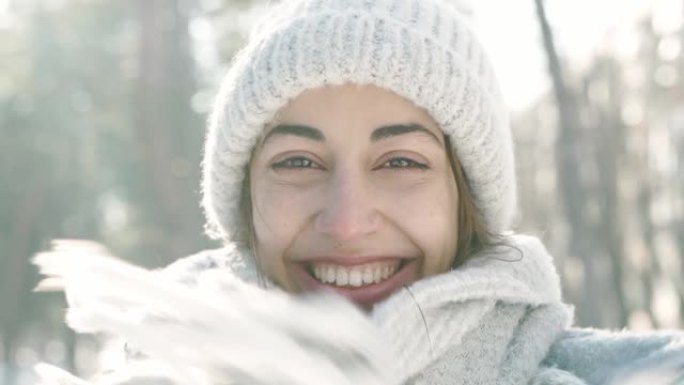 在寒冷的晴天，白雪皑皑的冬季公园里，穿着温暖的羊毛帽和长围巾的美丽大笑年轻女子的慢动作特写肖像。女人