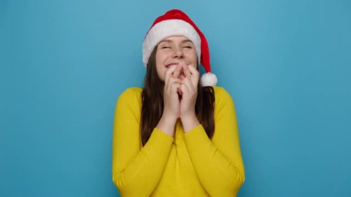 制作圣诞愿望概念。快乐可爱的年轻女子交叉手指站着，等待奇迹发生，戴着红色帽子和黄色毛衣，相信好运，孤