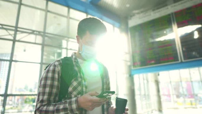 戴防护面罩和杯咖啡的男子使用智能手机在线办理登机手续，站在机场航站楼附近的出发到达时间表屏幕显示期间