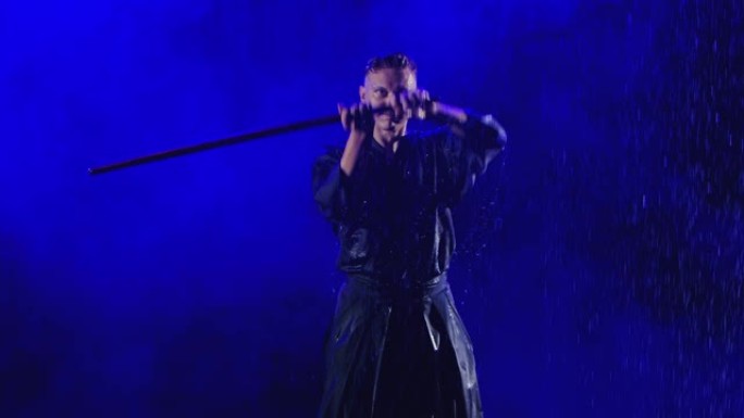 在雨水下，用蓝色和服的剑道大师的木剑锻炼。在黑烟背景下用蓝光慢动作拍摄。特写