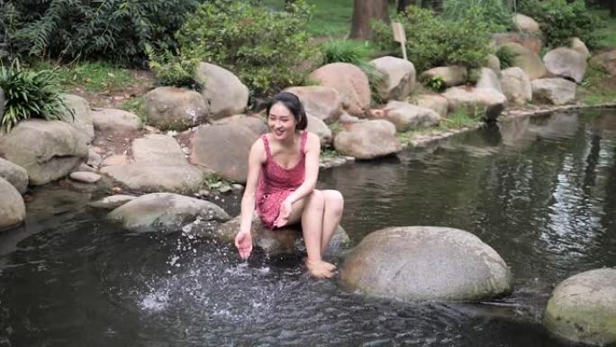 穿着红色连衣裙的性感中国年轻女子坐在河边的大石头上玩水，赤脚女孩在夏天享受水，超级慢动作镜头，夏天的
