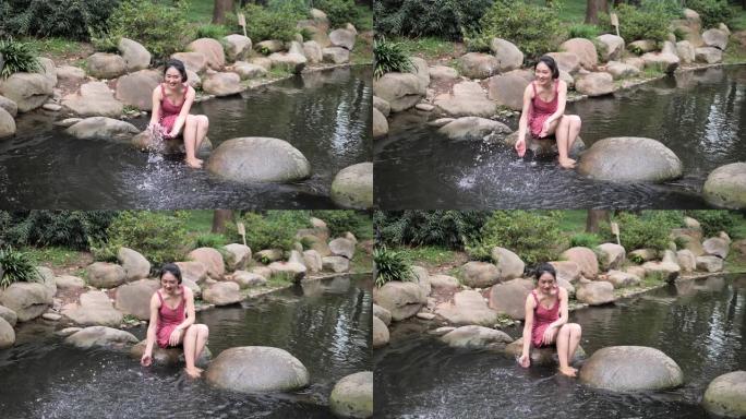 穿着红色连衣裙的性感中国年轻女子坐在河边的大石头上玩水，赤脚女孩在夏天享受水，超级慢动作镜头，夏天的