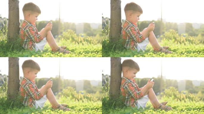 夏天日落时，坐在草地上的开朗男孩在手机里看着卡通。可爱的宝宝在大自然中玩得开心