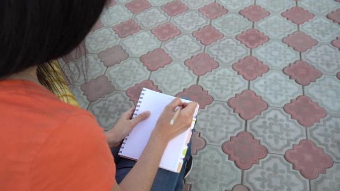 戴着医用口罩的女孩在日记中写下待办事项清单。社交远程，远程教育。学生正在公园里准备上课。一头黑发的女