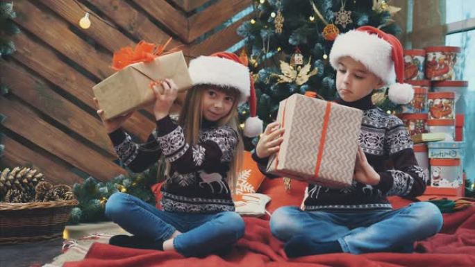 小双胞胎坐在圣诞树下，摇晃着摆弄着礼物的盒子，找出里面的东西。