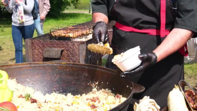一个穿着黑色衣服和手套的厨师把芬芳的抓饭放在一个盘子里，放在一个巨大的大锅里的街头食物上。街头节日新
