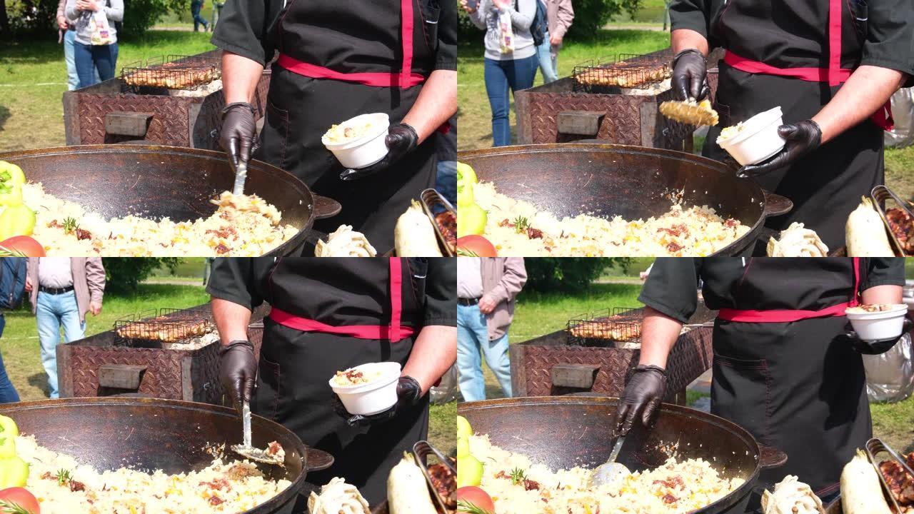 一个穿着黑色衣服和手套的厨师把芬芳的抓饭放在一个盘子里，放在一个巨大的大锅里的街头食物上。街头节日新
