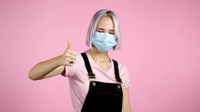 可爱的女人，戴着医用面具和紫色染色的发型，在粉色背景上显示拇指向上签名。积极的年轻女孩对镜头微笑。赢