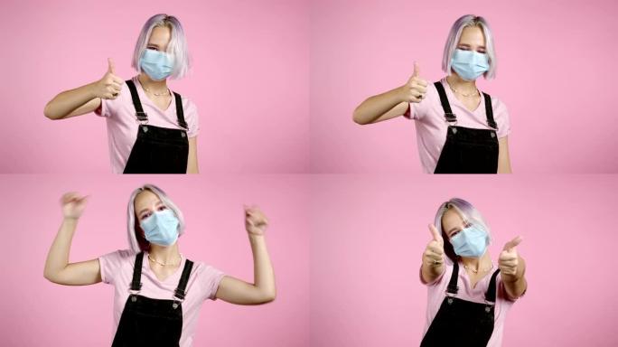 可爱的女人，戴着医用面具和紫色染色的发型，在粉色背景上显示拇指向上签名。积极的年轻女孩对镜头微笑。赢