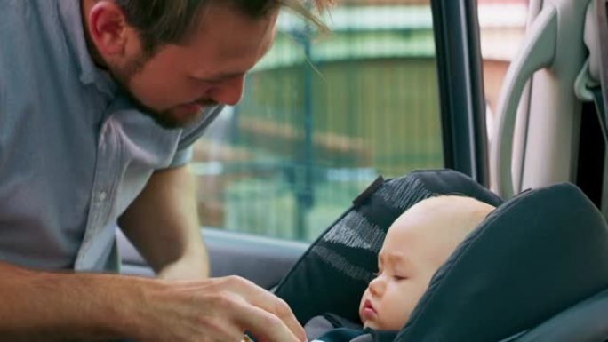 车内摄像头。特写男婴坐在车内的婴儿汽车座椅上。年轻的大胡子父亲检查婴儿系好的安全带，微笑并与他交谈，