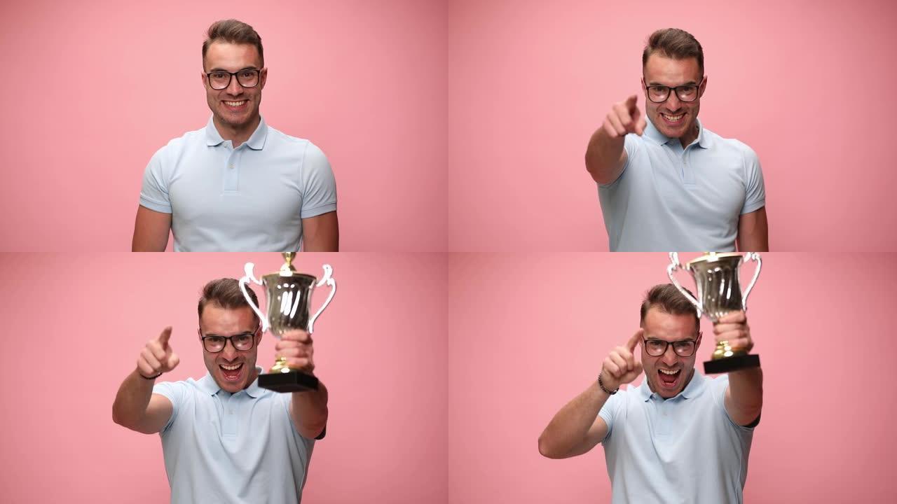 性感的休闲男人面带微笑，指着相机，举起奖杯，在粉色背景上庆祝成功
