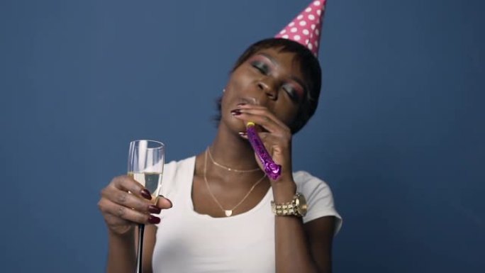 穿着生日帽的迷人非洲女人拿着香槟玻璃，蓝色背景上戴着生日喇叭吹口哨