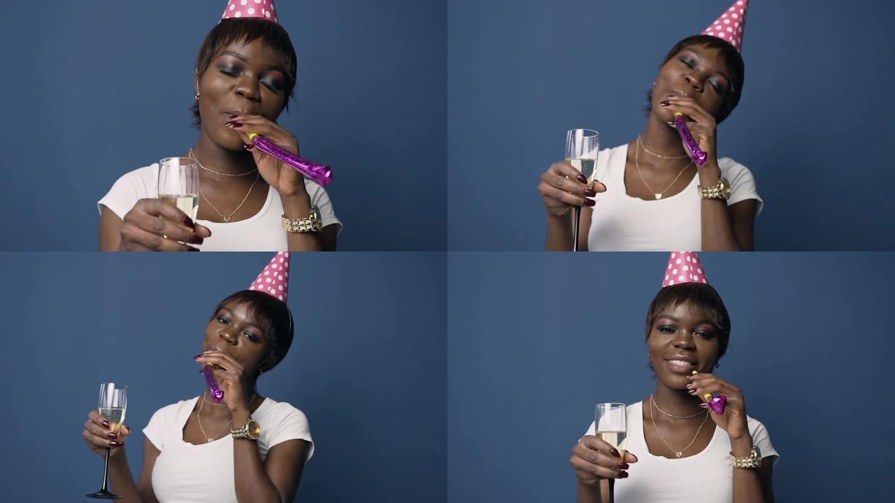 穿着生日帽的迷人非洲女人拿着香槟玻璃，蓝色背景上戴着生日喇叭吹口哨