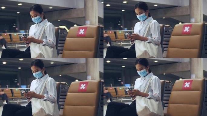 年轻的女商人戴着外科口罩，坐在机场候机楼的椅子上，并在机场使用手机登机。
