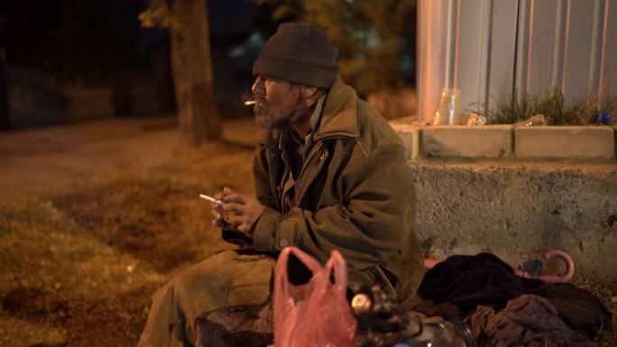 孤独寒冷的无家可归的人晚上坐在街上抽烟。