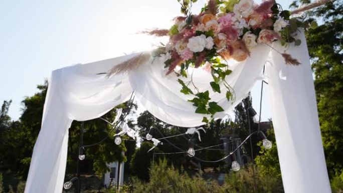 阳光明媚的夏日用鲜花装饰的婚礼拱门。节日花卉装饰。玫瑰和羽毛草，白色纺织框架上有灯泡花环
