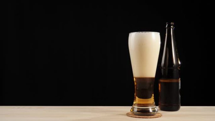 新鲜啤酒。手将一个棕色的瓶子和美味的精酿啤酒放在一杯啤酒旁边，放在黑色背景下的木制桌子上。带有水滴，