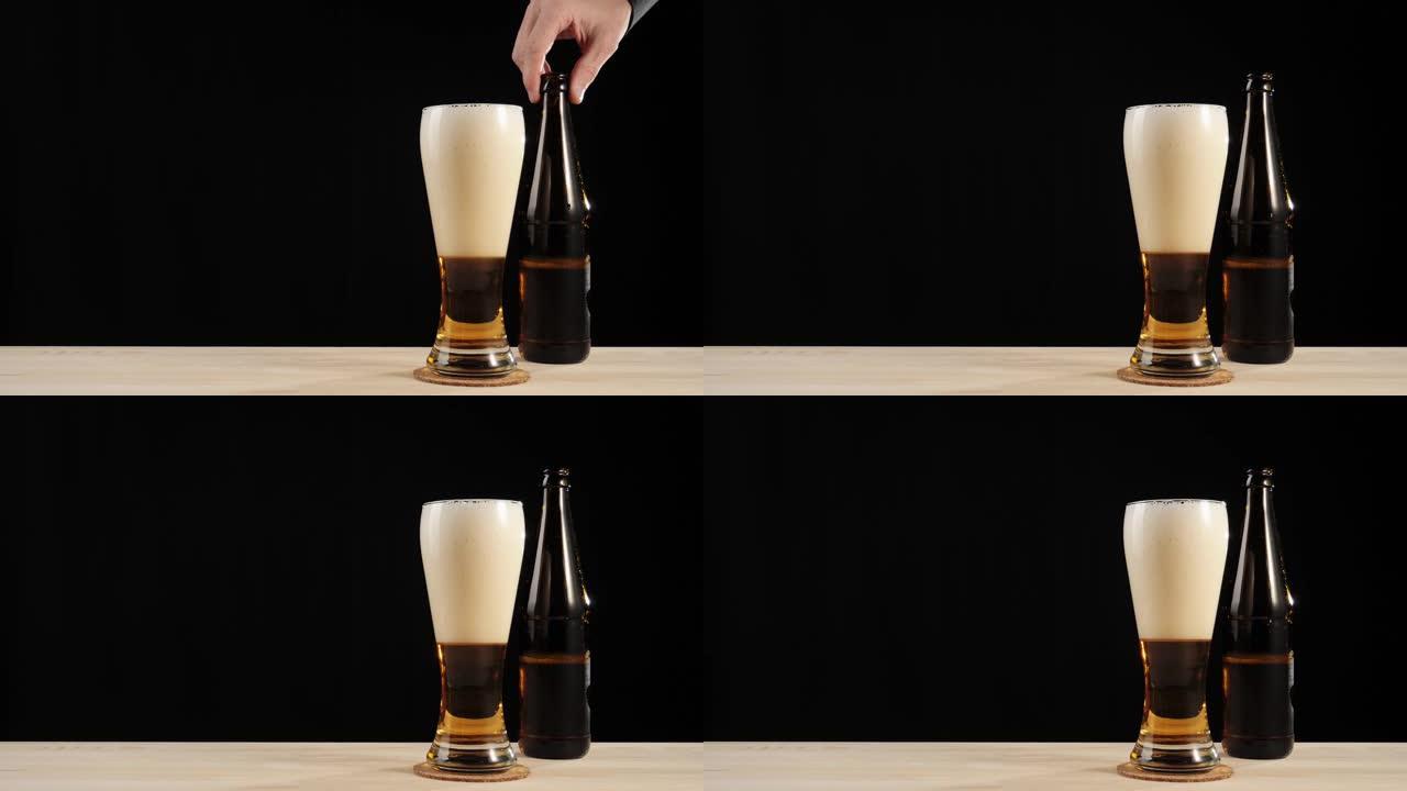 新鲜啤酒。手将一个棕色的瓶子和美味的精酿啤酒放在一杯啤酒旁边，放在黑色背景下的木制桌子上。带有水滴，