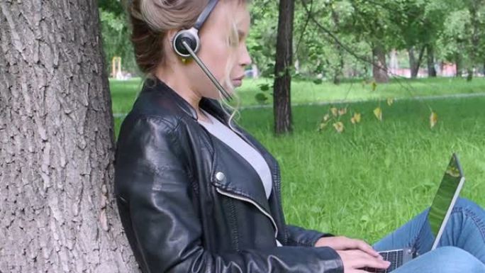 微笑的女人坐在城市公园里，戴着笔记本电脑和耳机进行在线对话。侧视图