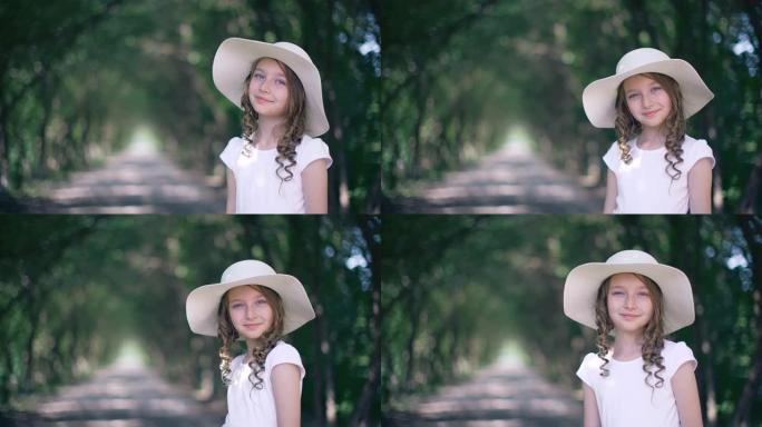 绿色公园背景下帽子卷发的肖像微笑女孩。戴着夏日帽子的蜂蜜卷毛女孩看着树巷走廊上的相机。