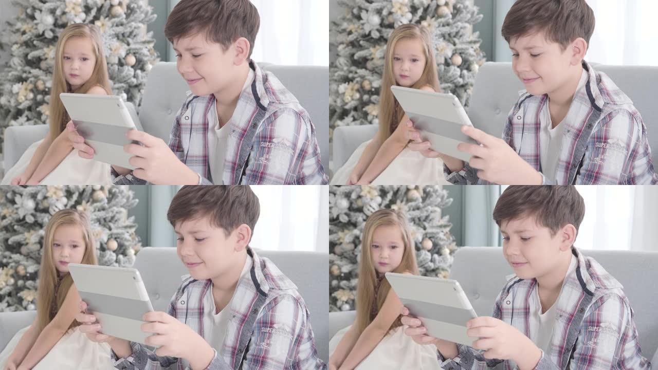 可爱的高加索女孩坐在背景上，看着男孩玩平板电脑的肖像。平安夜，穿着漂亮裙子的害羞小女士和黑发男孩躺在