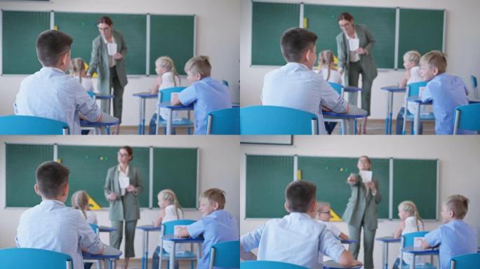 课上课桌上的同学互相交谈，老师在黑板附近给学生看卡片，并问学校里的男孩，后视图
