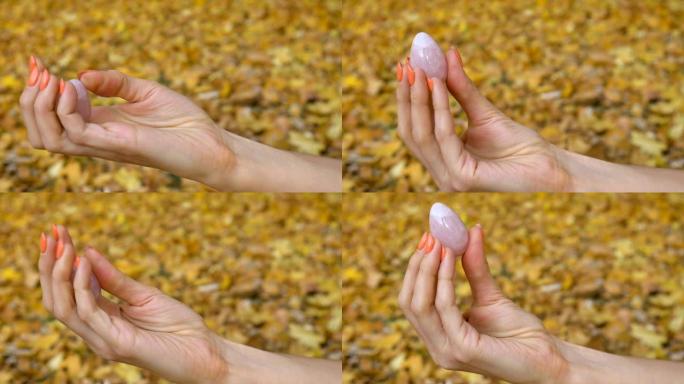 橙色修指甲的女性手拿着粉色石英约尼蛋，适合秋季户外黄色落叶背景的vumfit、imbuilding或