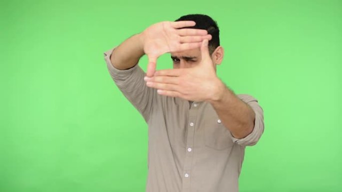 布鲁内特男子用手指透过框架形状，用专业摄影师的目光聚焦相机，捕捉照片。绿色背景，色度键