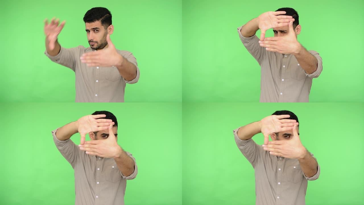 布鲁内特男子用手指透过框架形状，用专业摄影师的目光聚焦相机，捕捉照片。绿色背景，色度键