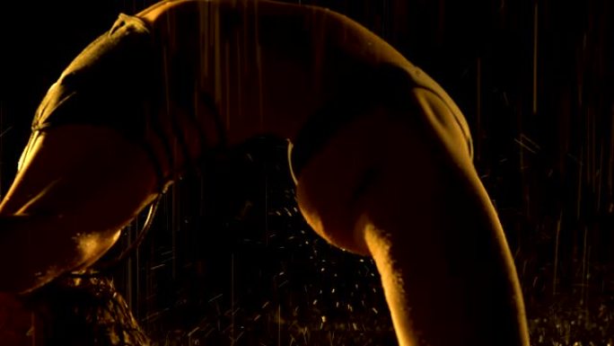 剪影被雨淋湿的苗条女人在黑暗的工作室里练习瑜伽体式。性感的身体覆盖着在工作室黄光下闪闪发光的水滴。黑